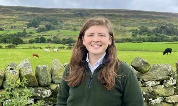 Headshot of a woman with long brown hair stood outside in front of a stone wall.
